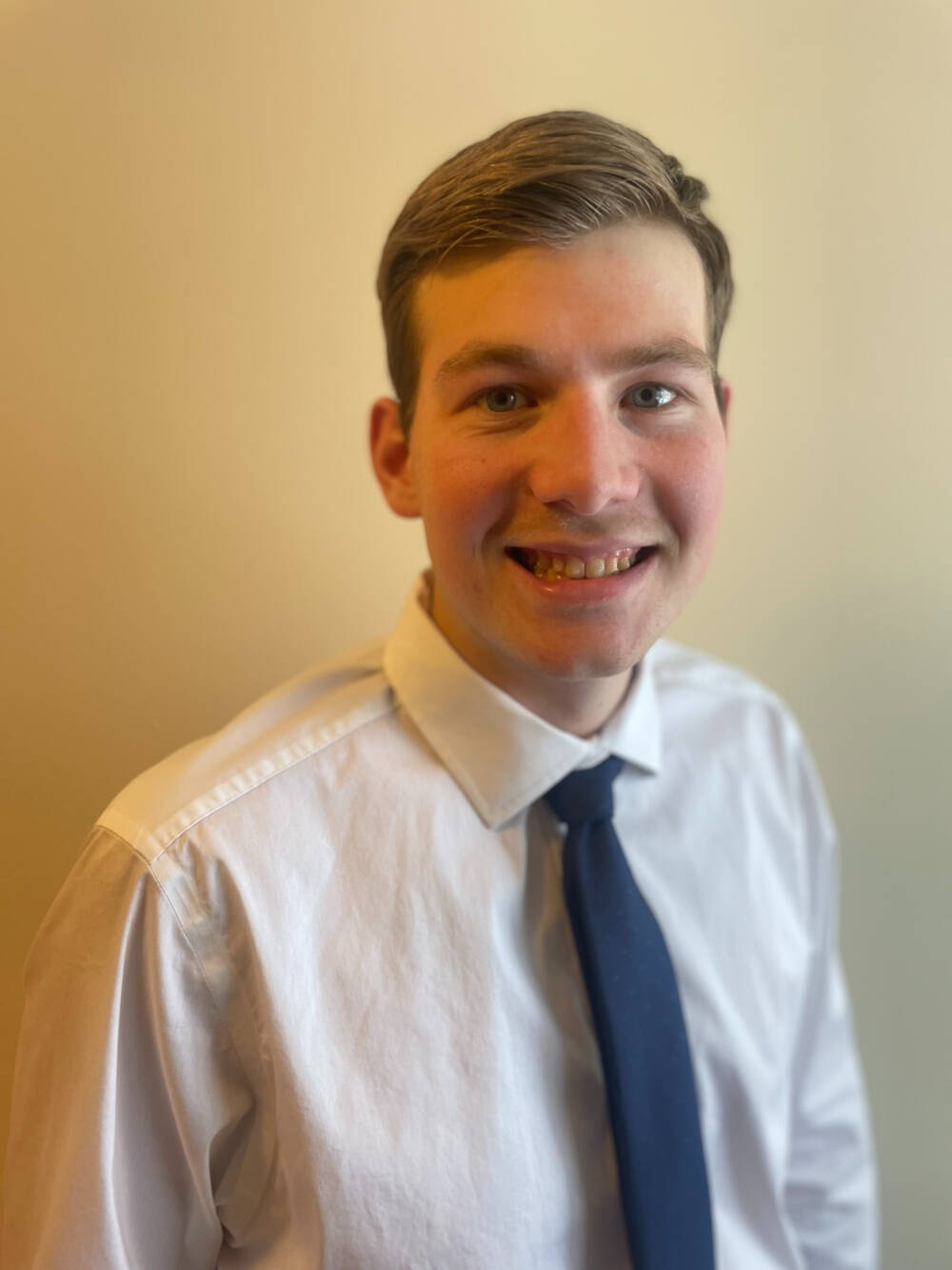 Colin Nagley, in a White official shirt and a blue tie smiles. Behind him is a nude brown background.